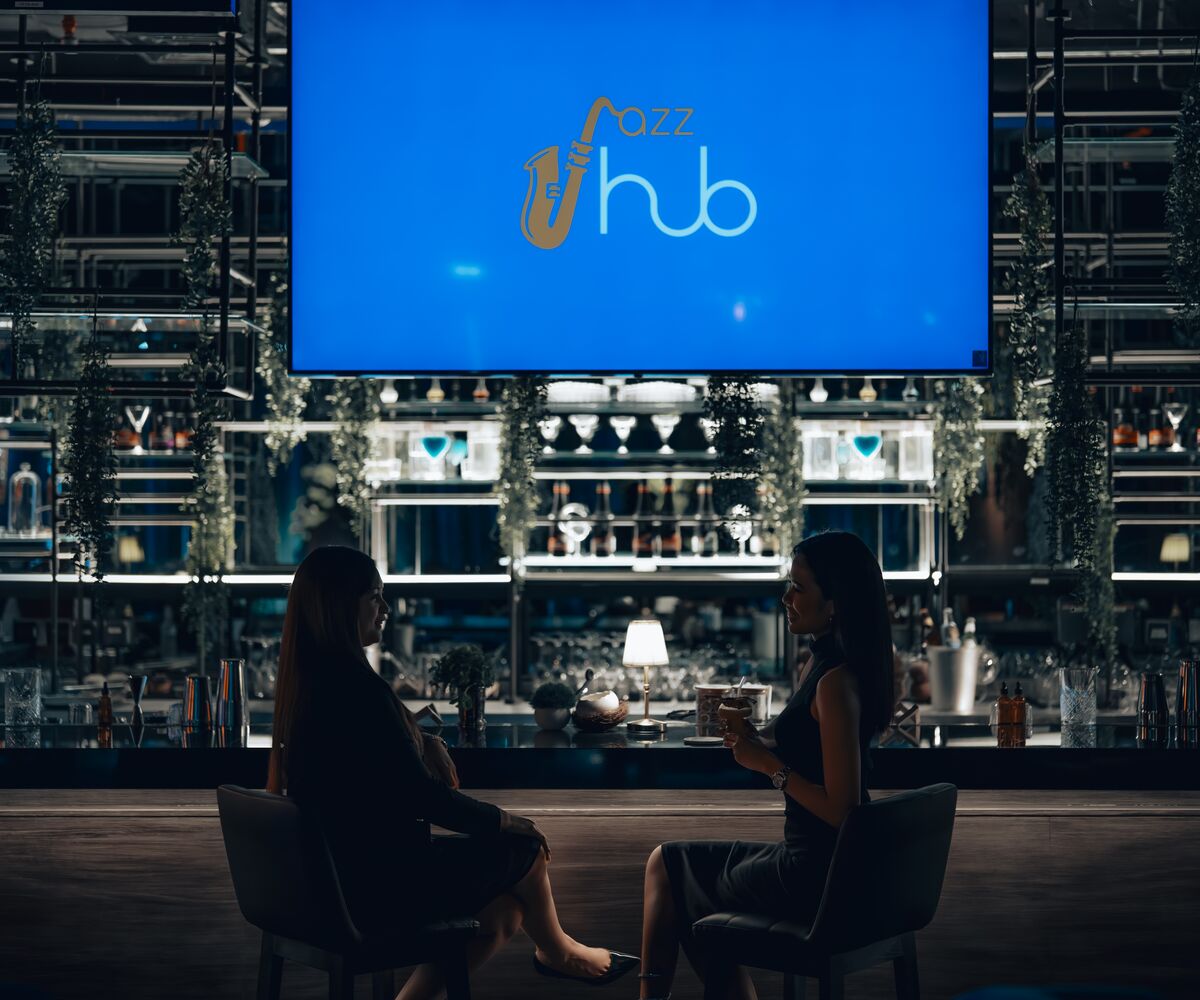 Two guests chatting at the Jazz Hub bar beneath a large screen showing the Jazz Hub logo